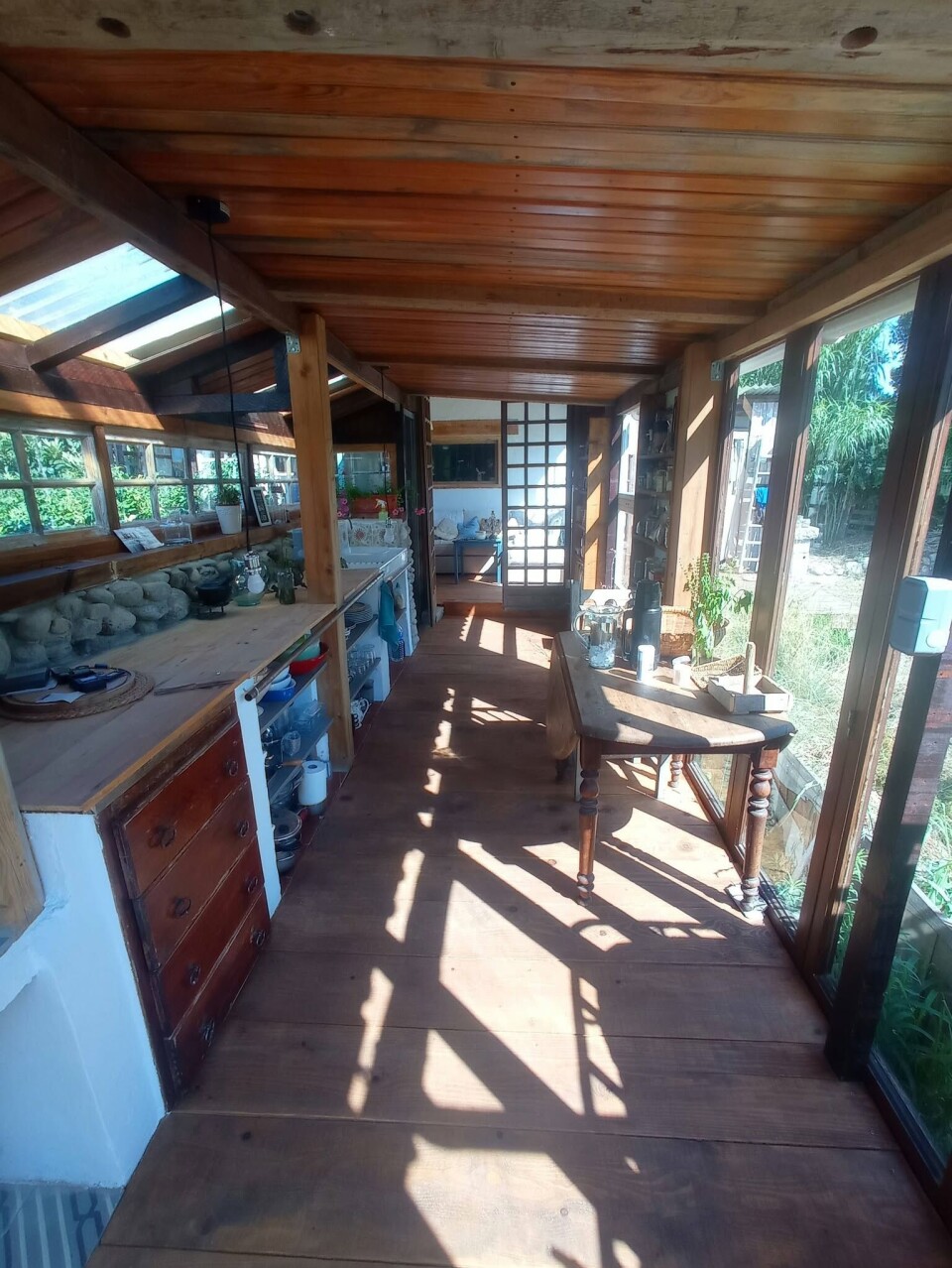 Sunlit rustic timber veranda with windows, plants, a table and kitchen fittings.